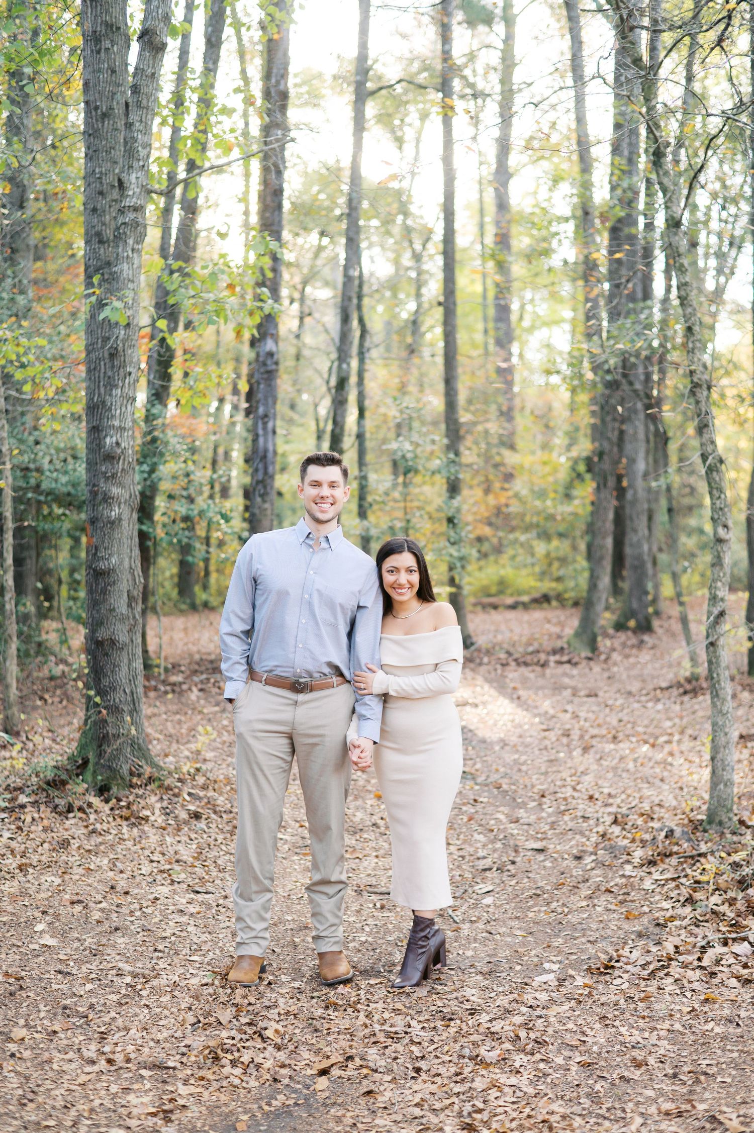 A couple stands together on a leaf-covered forest path during autumn with tall trees surrounding them.