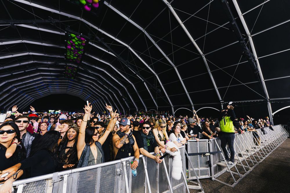 Crowd of concert-goers raise their hands in excitement inside a dark venue with neon lighting and curved ceiling.