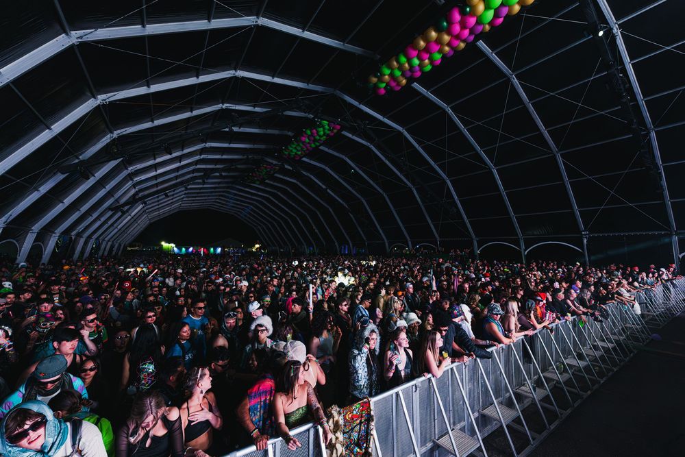 Concert crowd stands in curved rows under dark lighting with colorful spotlights illuminating the scene.