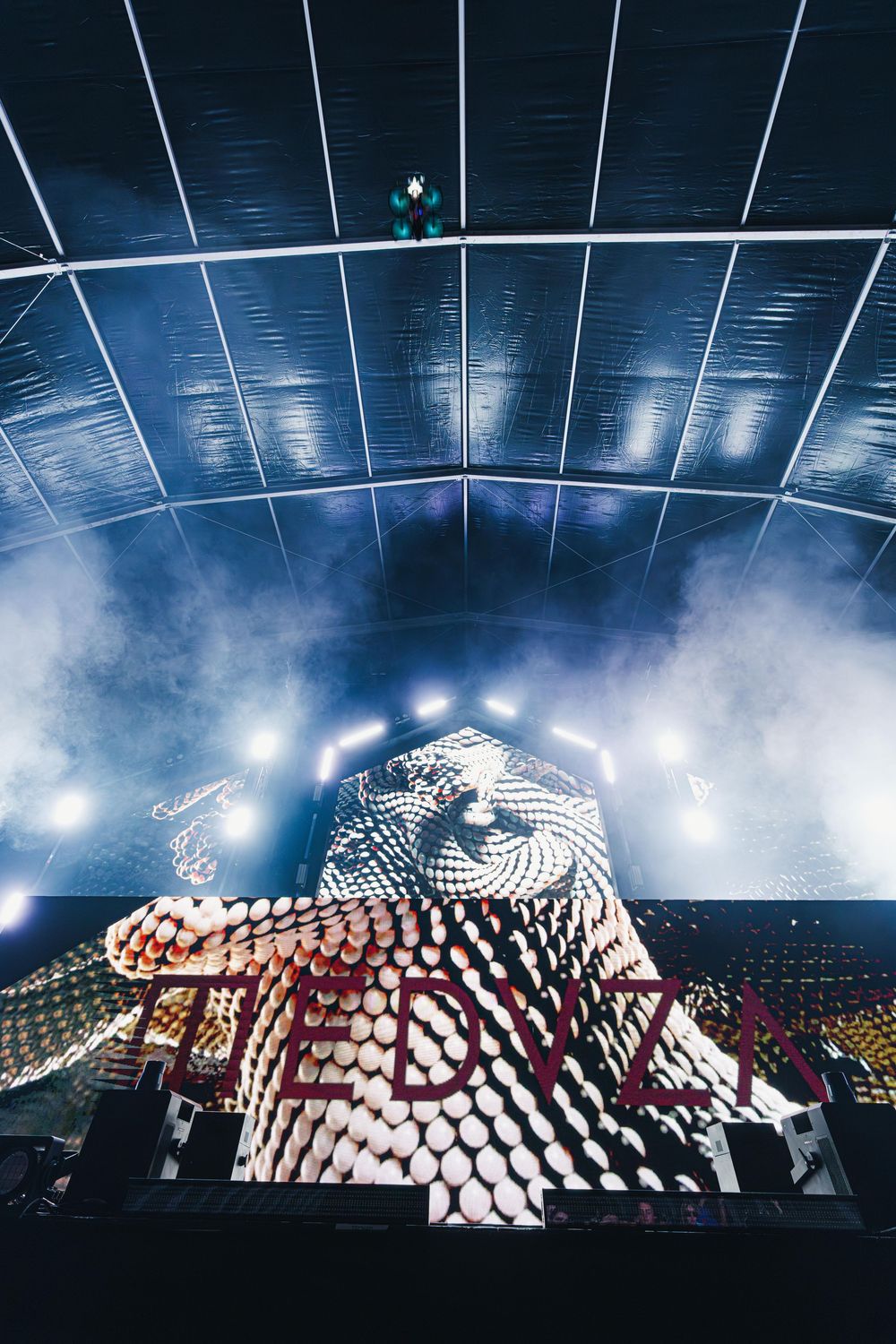 A large geometric stage design with dramatic lighting and fog effects creates an atmospheric concert environment.