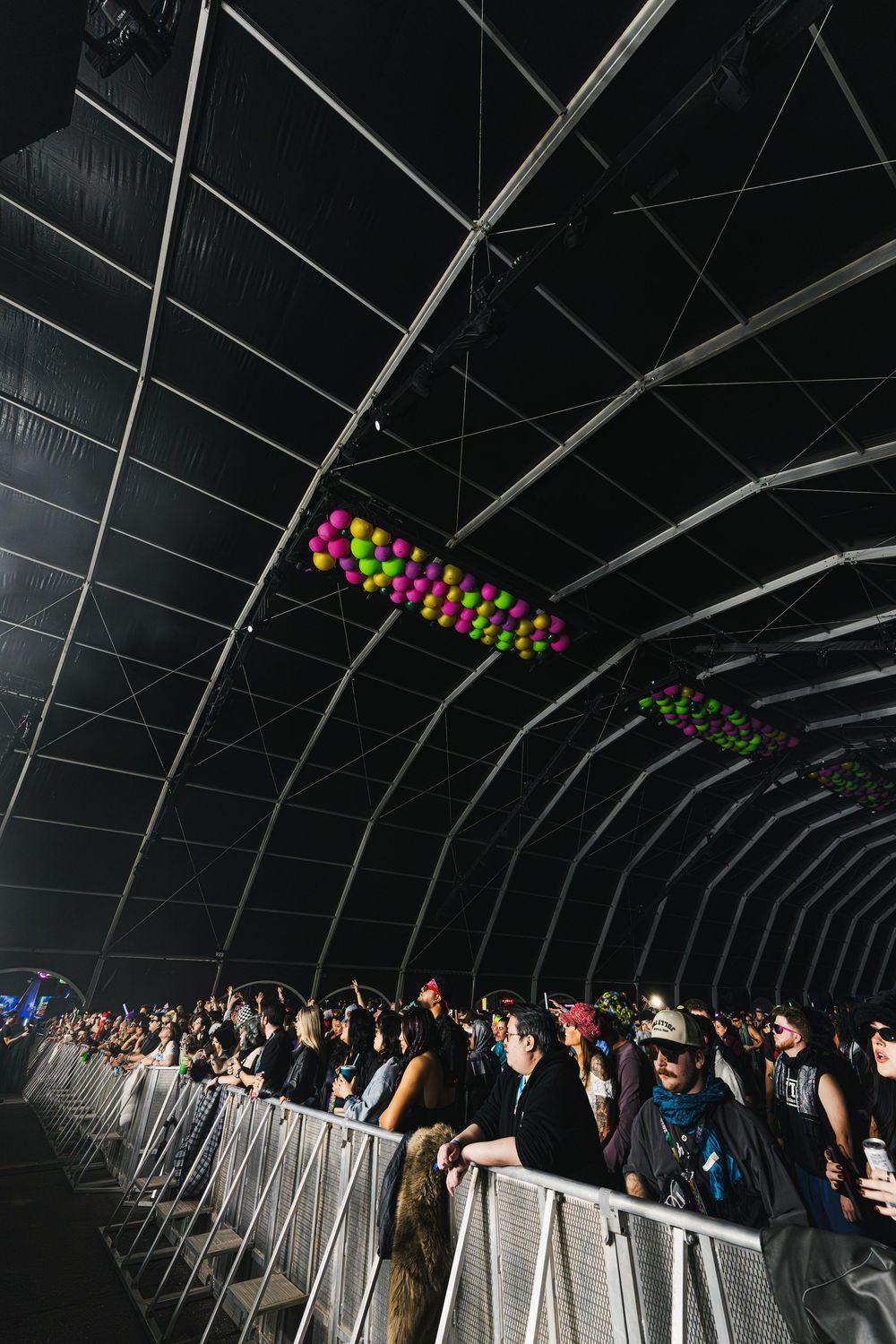 A crowd of people lined up against a barrier while colorful balloons float above in a dark tunnel-like space.
