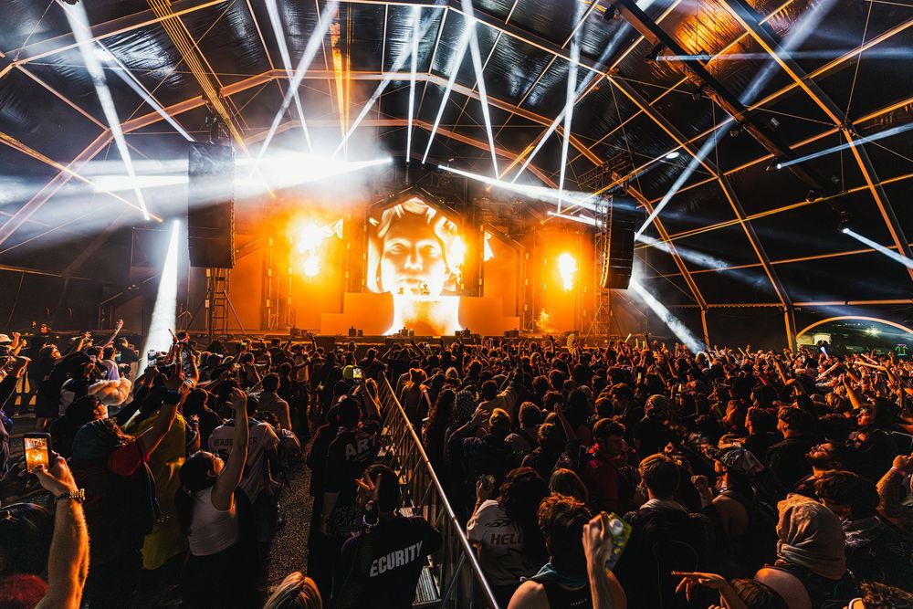 Concert crowd illuminated by bright stage lights and beams during nighttime performance at large indoor venue.