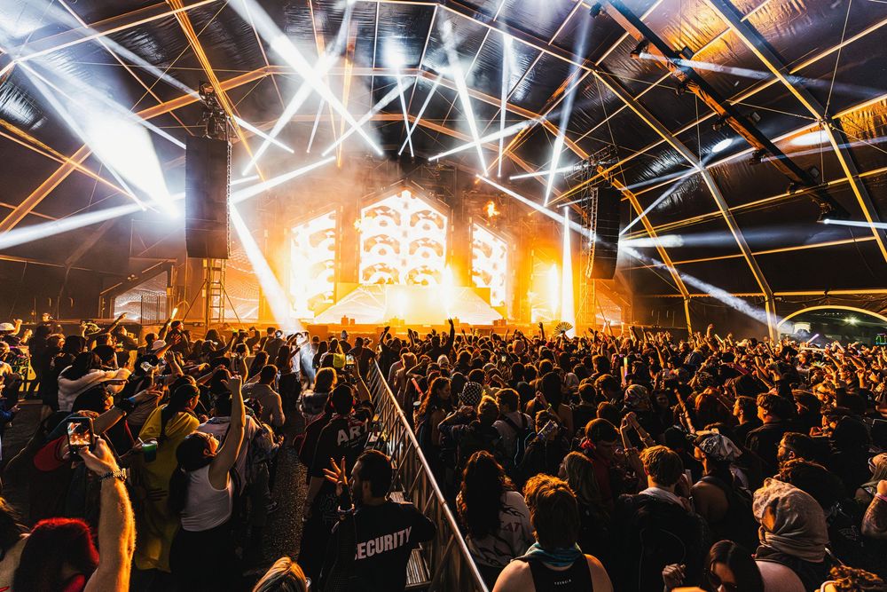 Large concert crowd in dark venue illuminated by bright stage lights and flashing effects during live performance.