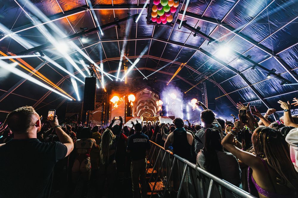 Concert crowd at night venue with colorful stage lights illuminating arched ceiling and energetic atmosphere.