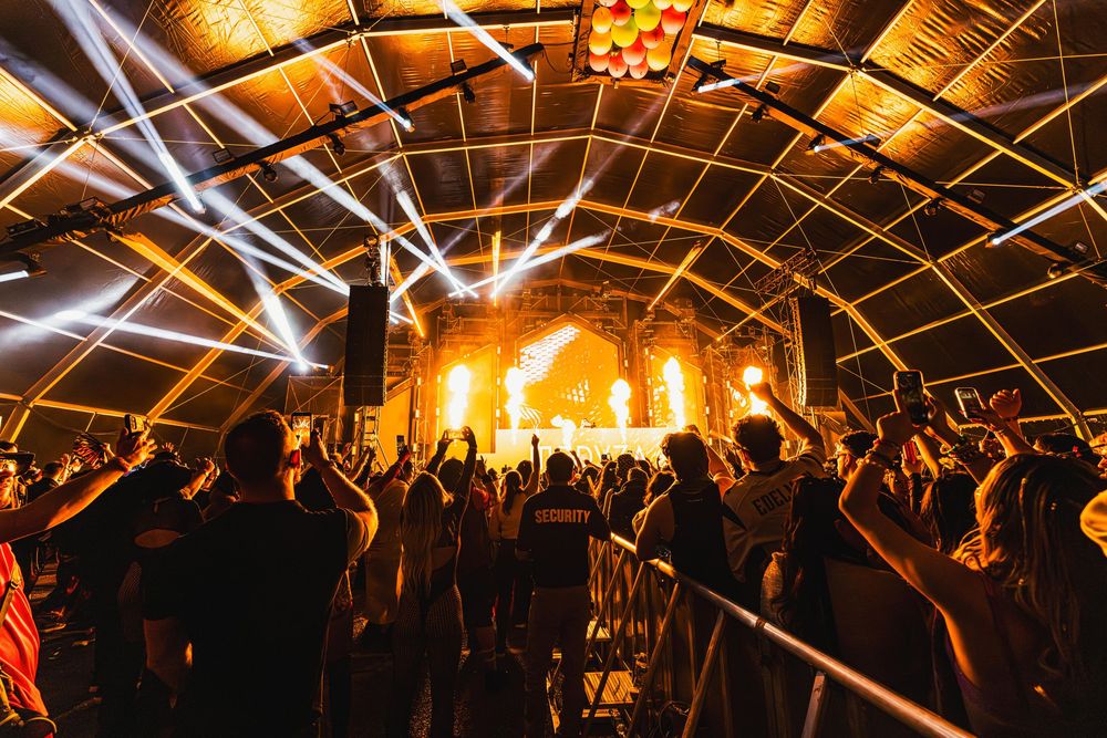 Concert crowd silhouettes in a warehouse venue with bright orange stage lights and illuminated ceiling beams.
