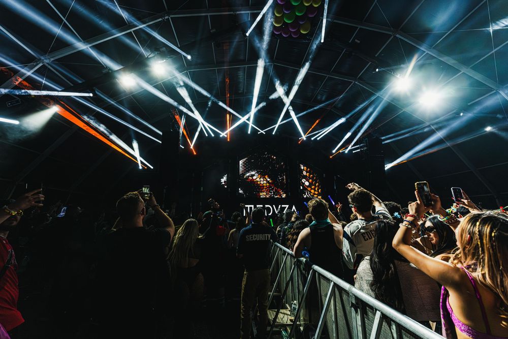 Concert crowd raises their hands in excitement as bright stage lights illuminate a dark venue during a live performance.