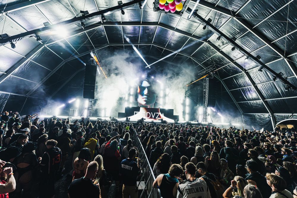 Large crowd of concert attendees filling a dark arena with dramatic overhead lighting and atmospheric smoke effects.