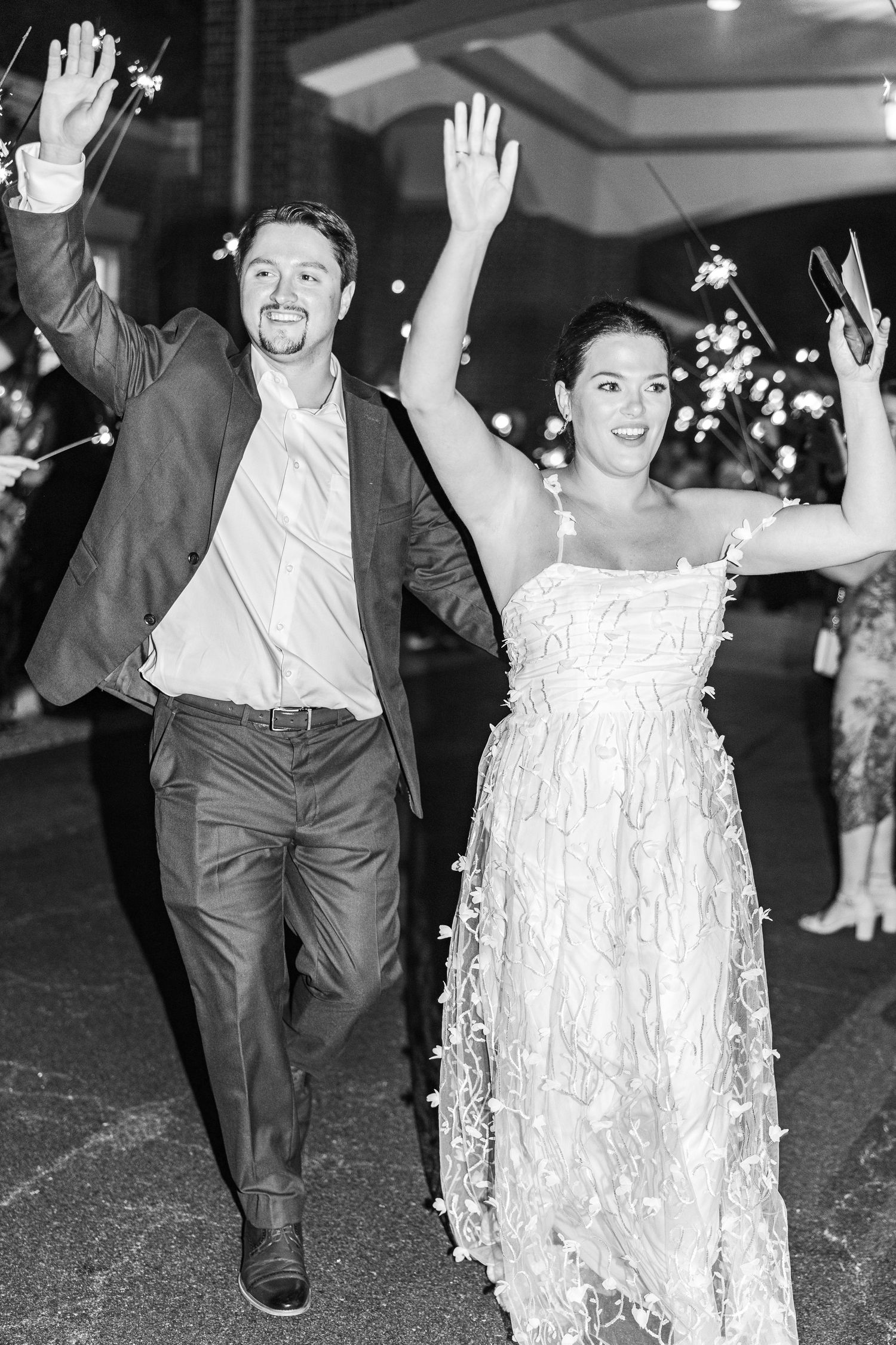 Black and white photo of newlyweds joyfully exiting their wedding reception as guests throw confetti and rice.