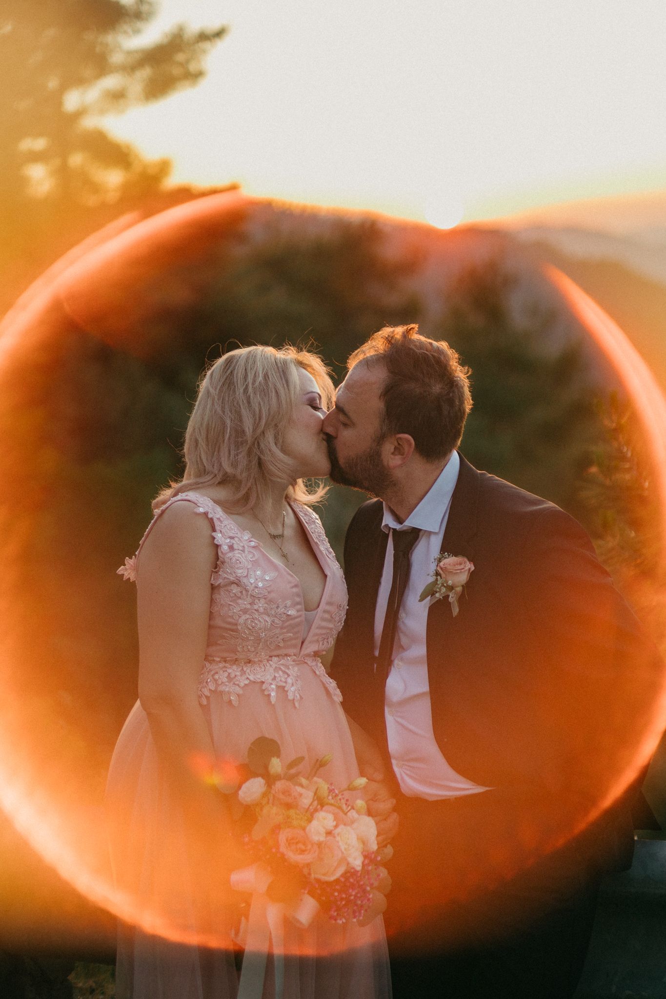 A romantic couple shares intimate moments at sunset with orange lens flares creating dreamy heart-shaped bokeh effects.
