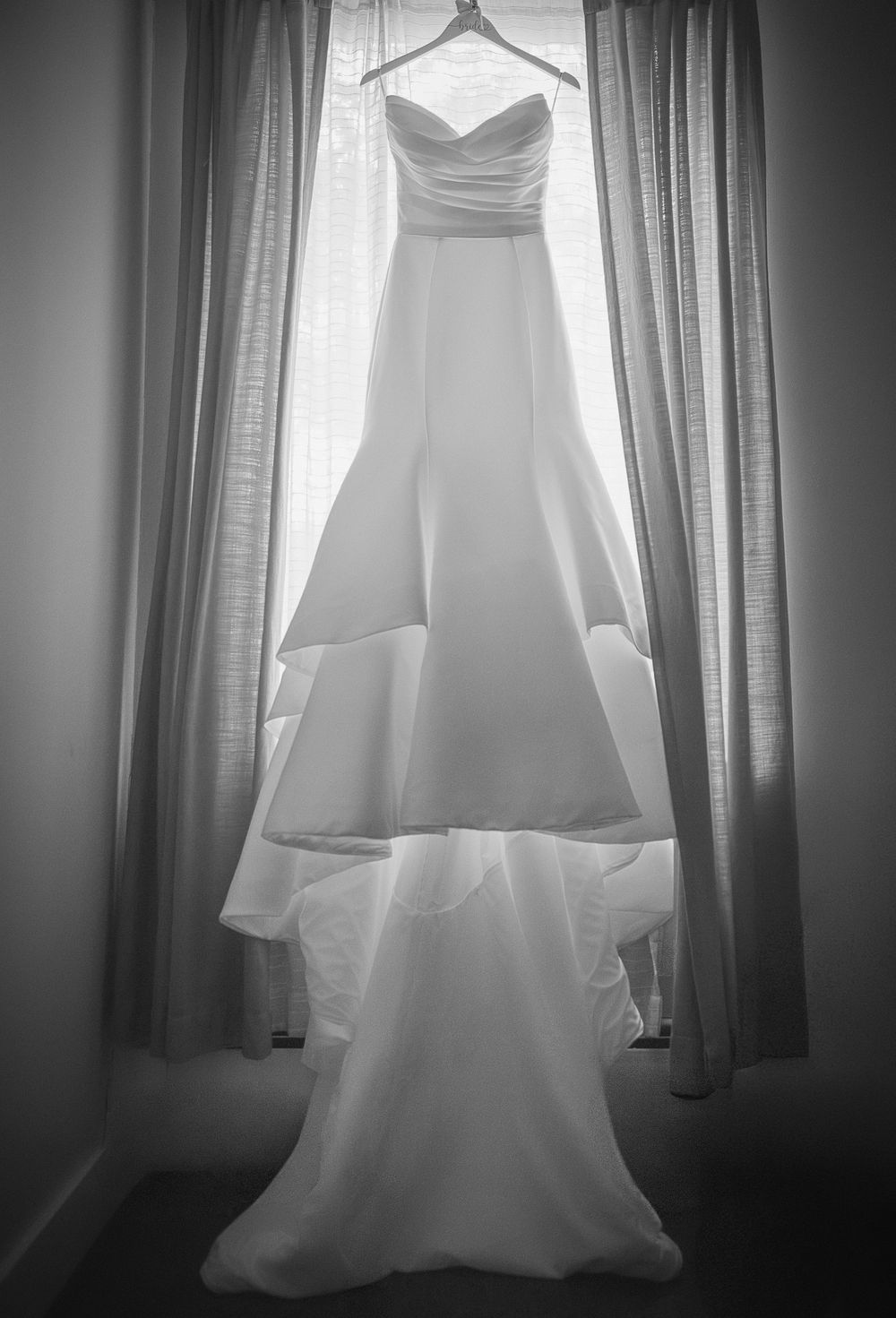 A white wedding dress hangs gracefully in front of a window with soft natural light filtering through.