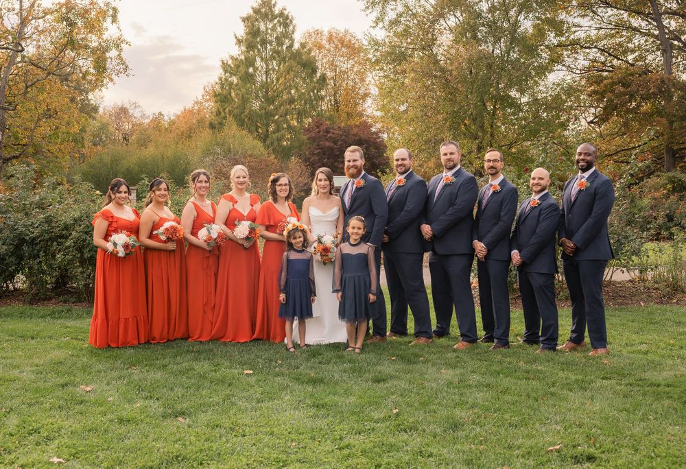 Wedding party in navy and coral attire poses on grassy hill during fall season. Park of Roses Columbus.