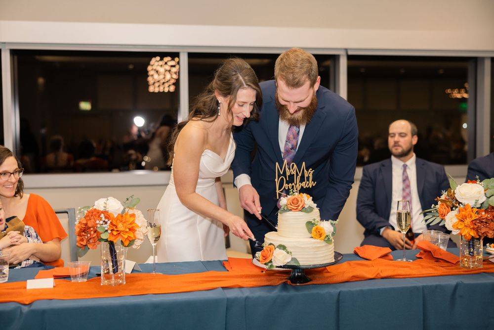 Wedding guests gather around a table decorated with orange accents and floral centerpieces at an evening reception. Bob crane event center