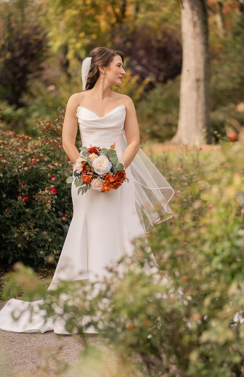 Bride in elegant white dress holds vibrant fall bouquet in garden setting.