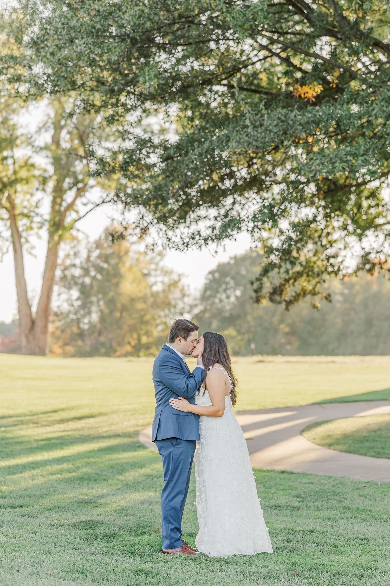 A couple shares a romantic moment on a grassy hillside during sunset, surrounded by trees in a park setting.