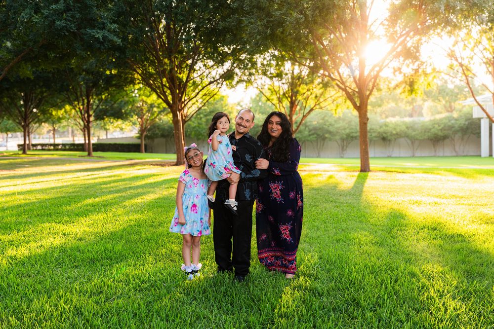 Family of four posing on lush green grass at golden hour with sunlit trees in background.