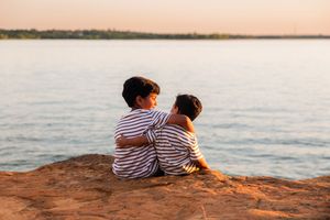 Two children in striped shirts sit embracing on a rocky cliff overlooking a calm lake at sunset. Murrell Park Flower Mound Lake Grapevine