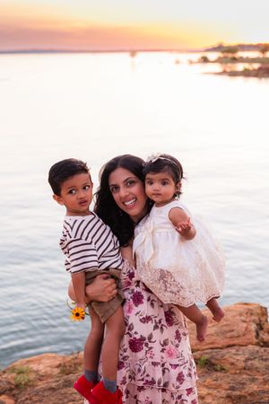 A smiling mother holds two young children at sunset by the water. Murrell Park Flower Mound Lake Grapevine