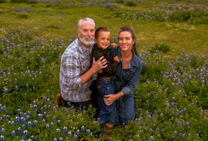 Family of three smiling together in a field of purple wildflowers on a sunny day. Family session irving texas bluebonnets