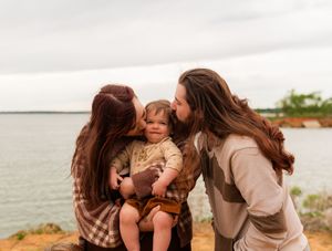 Two parents with long hair kissing their toddler on the cheeks outdoors near a scenic waterfront. Murrell Park Flower Mound Lake Grapevine