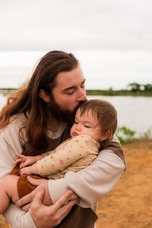 A bearded man with long hair tenderly kisses a sleepy baby outdoors near a lake. Murrell Park Flower Mound Lake Grapevine
