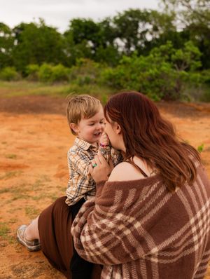 A mother and young boy share a joyful moment sitting on red dirt ground. Murrell Park Flower Mound Lake Grapevine