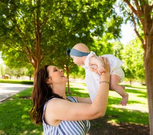 A smiling mother lifts her baby girl in a sunny park surrounded by lush green trees. Kimbell Art Museum Fort Worth Texas Family Session
