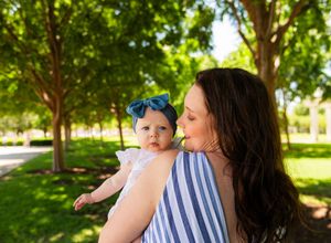 A mother holds her baby girl wearing a blue bow headband in a sunny green park. Kimbell Art Museum Fort Worth Texas Family Session with baby