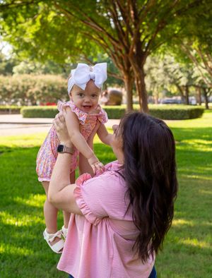 A woman in pink holds and plays with a baby wearing white in a sunny park setting. family session kimbell art museum fort worth texas