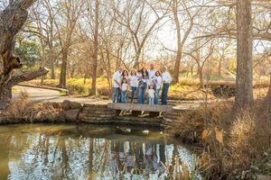 A group of people stand along a scenic pond surrounded by bare winter trees in a tranquil park setting.