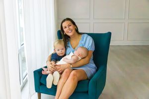 A parent and child sit together in a teal armchair next to a bright window in a white-walled room.