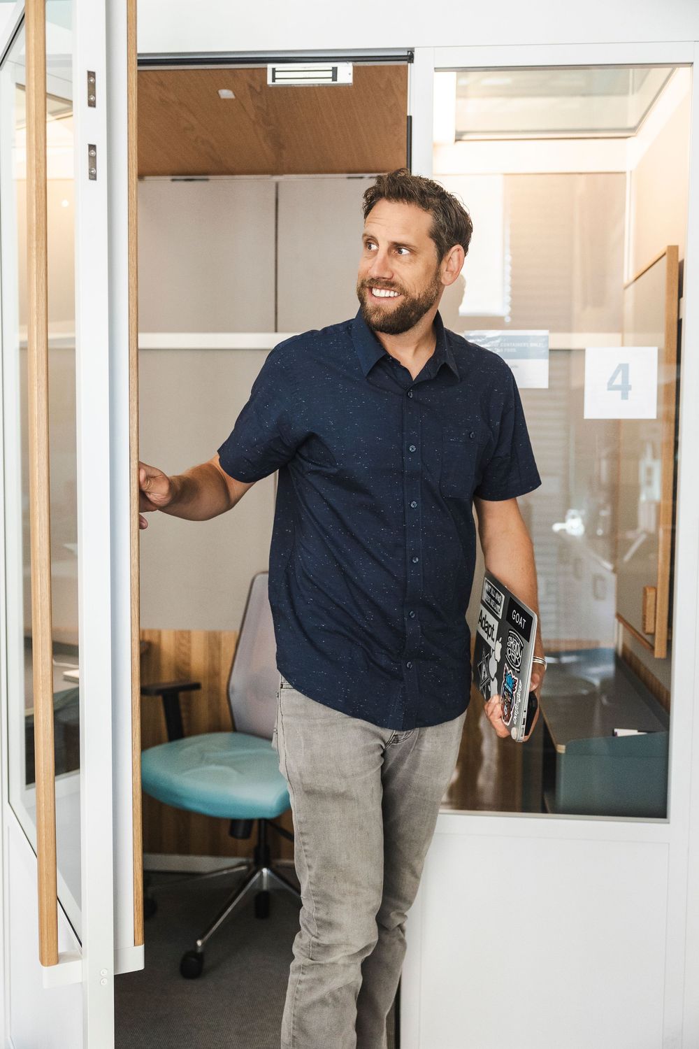 A person in a navy blue shirt and gray pants stands in a modern office hallway with glass walls and doors.
