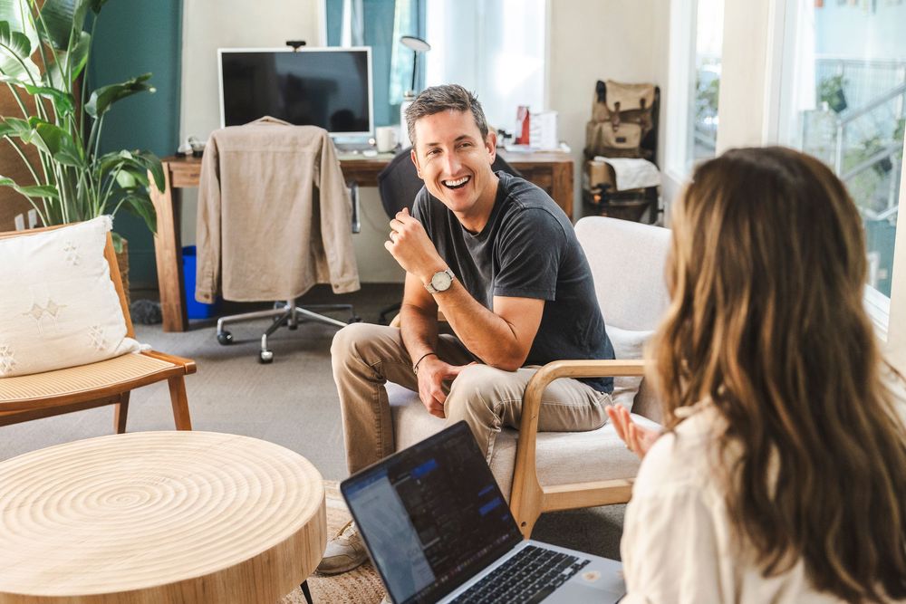 People having an engaging conversation in a bright modern living room with houseplants and natural lighting.