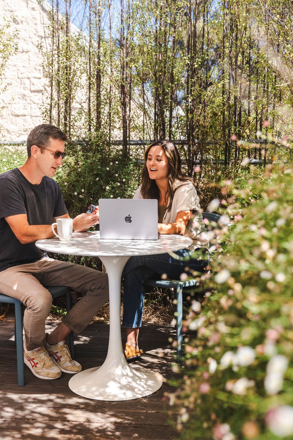 Two people having coffee and working on a laptop at an outdoor cafe table surrounded by blooming flowers.