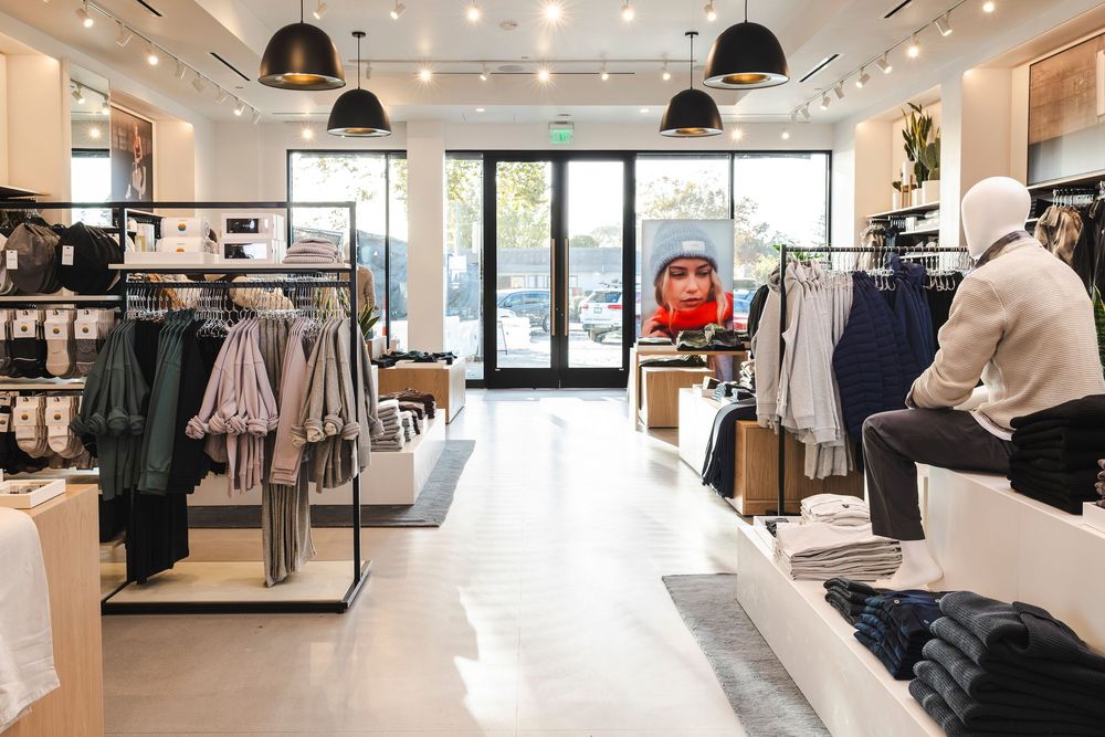 Modern clothing retail store interior with racks of apparel, pendant lighting, and glass entrance doors.