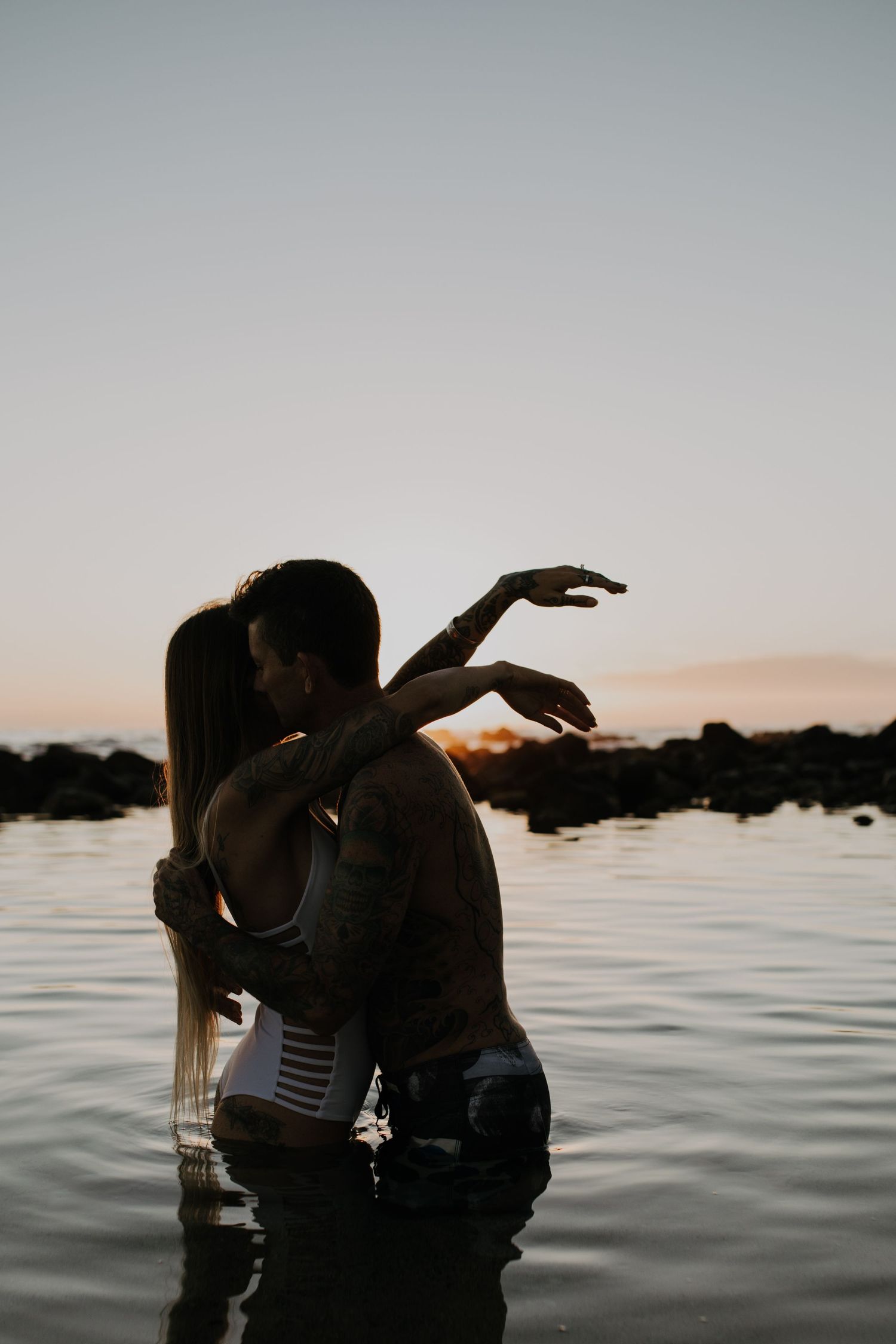 Silhouetted figures share an intimate moment in the water during sunset with a misty ocean backdrop.