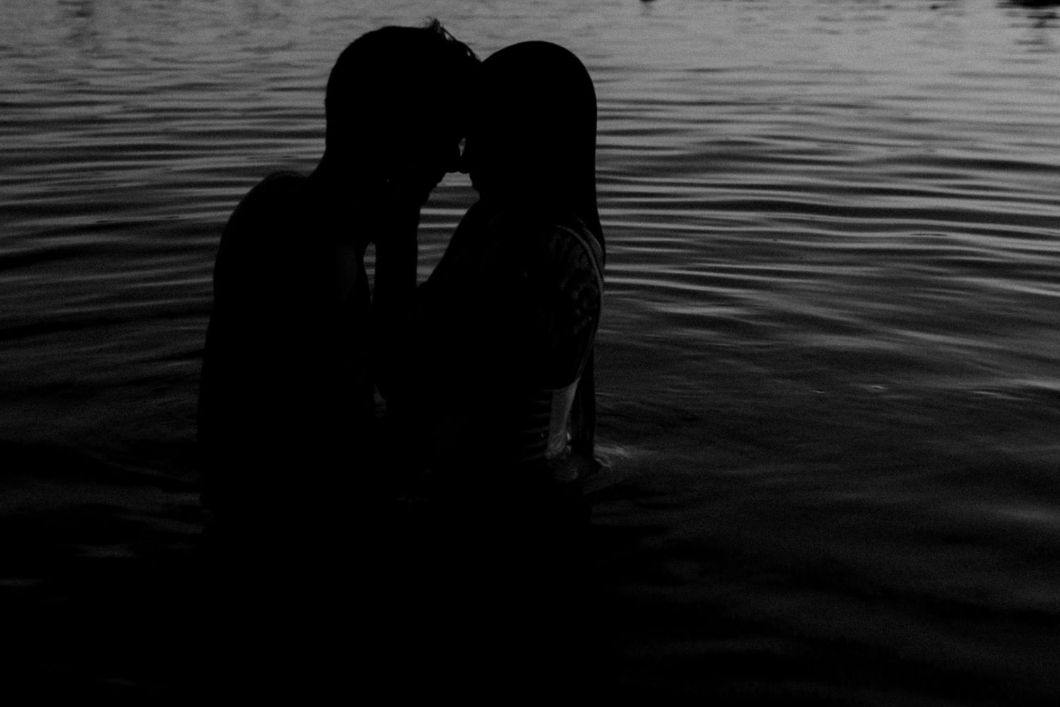 Silhouettes of people standing by calm water at sunset create a peaceful waterfront scene.