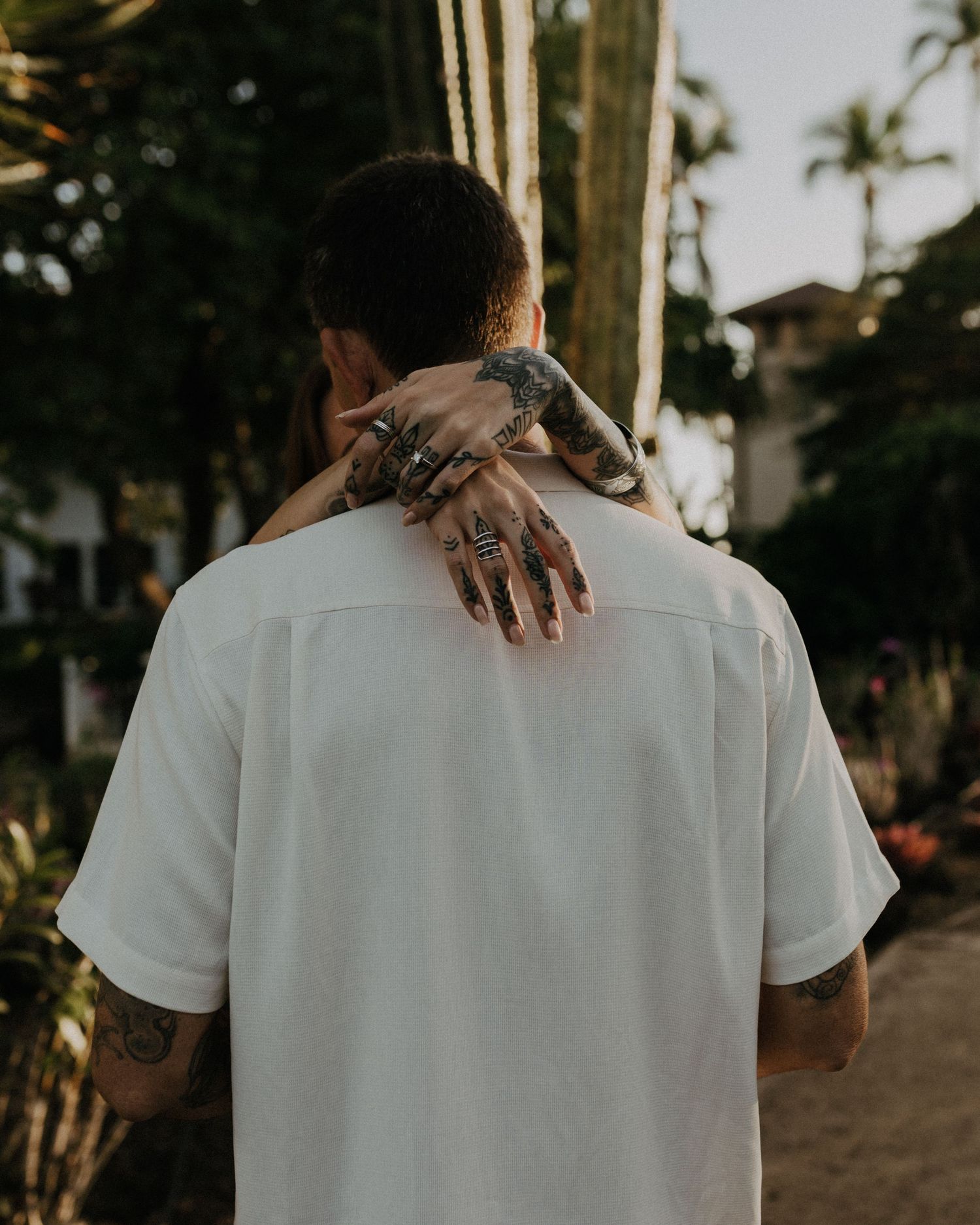 A couple embraces from behind in white clothing against a palm tree backdrop at sunset.