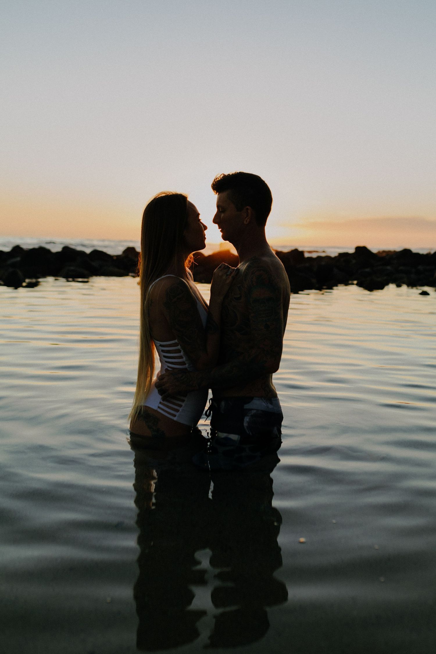 Silhouetted couple embraces in calm ocean waters during golden sunset.
