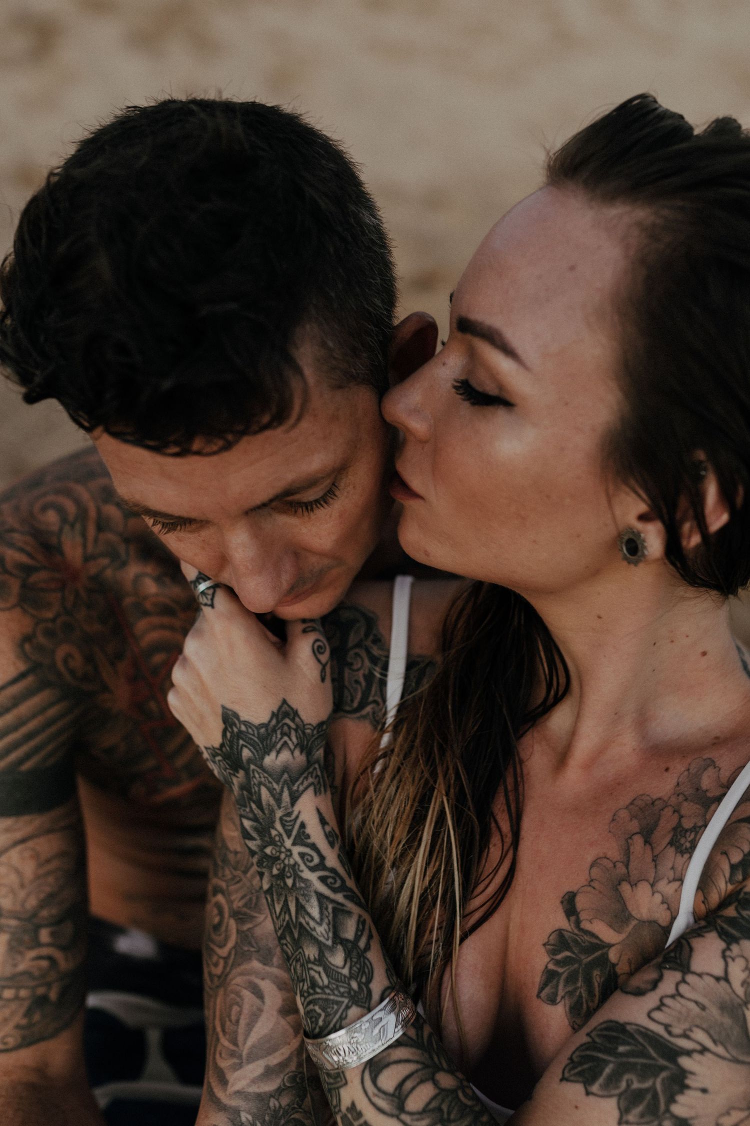 A romantic close-up portrait of a tattooed couple sharing an intimate moment against a neutral background.