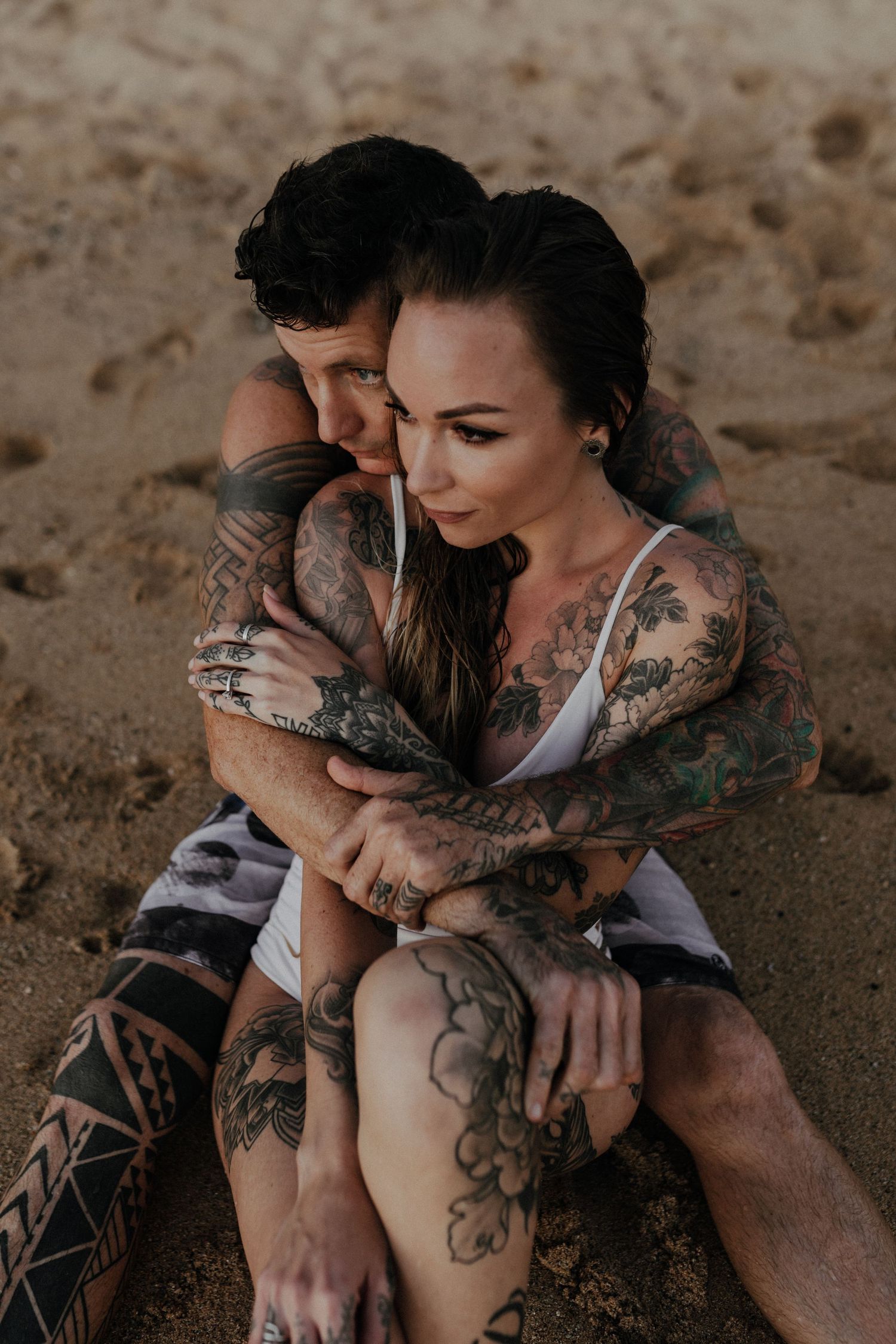 Tattooed couple sharing intimate moments on sandy beach during sunset photoshoot.