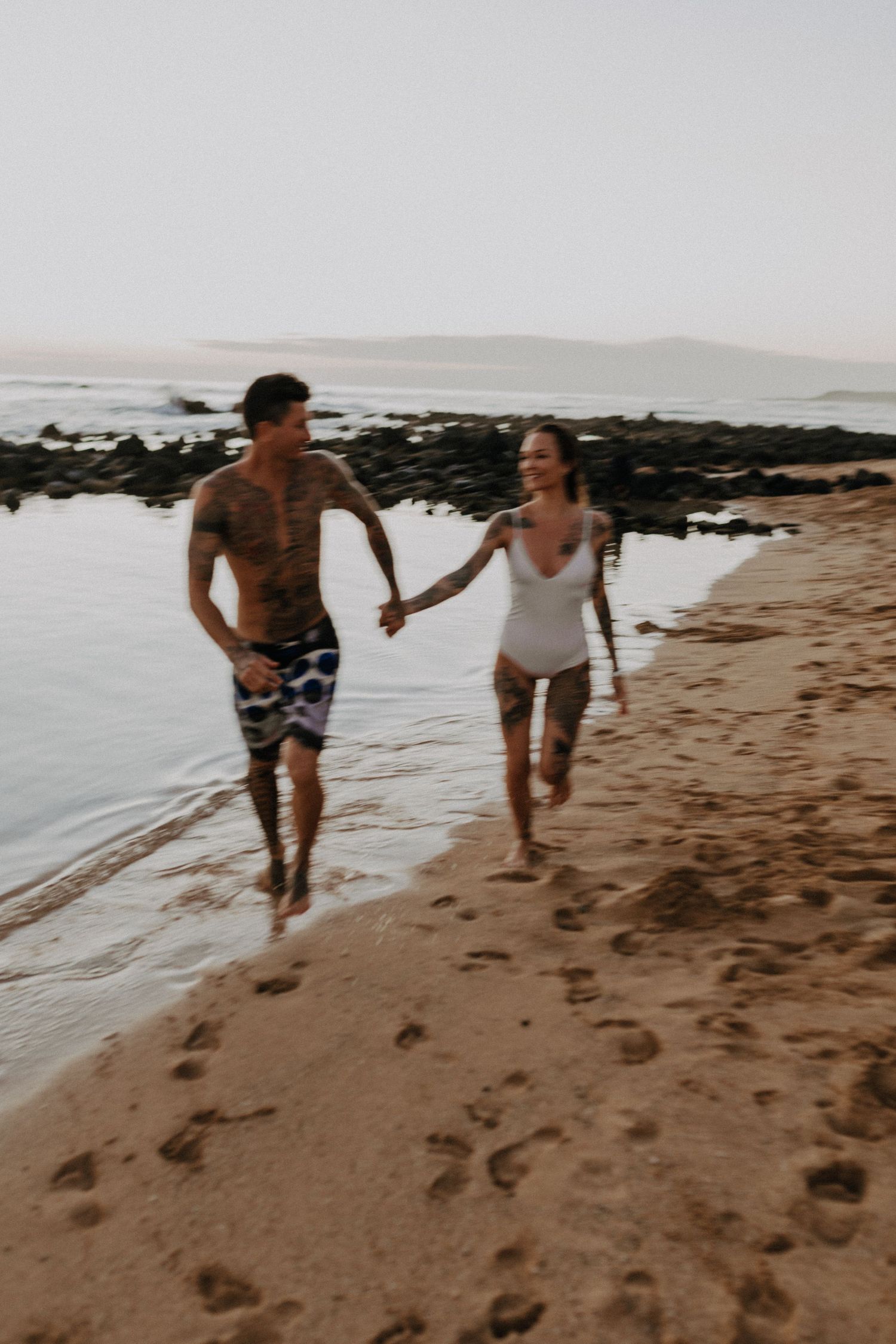 Two people run and laugh together along a sandy beach at sunset with ocean waves in the background.