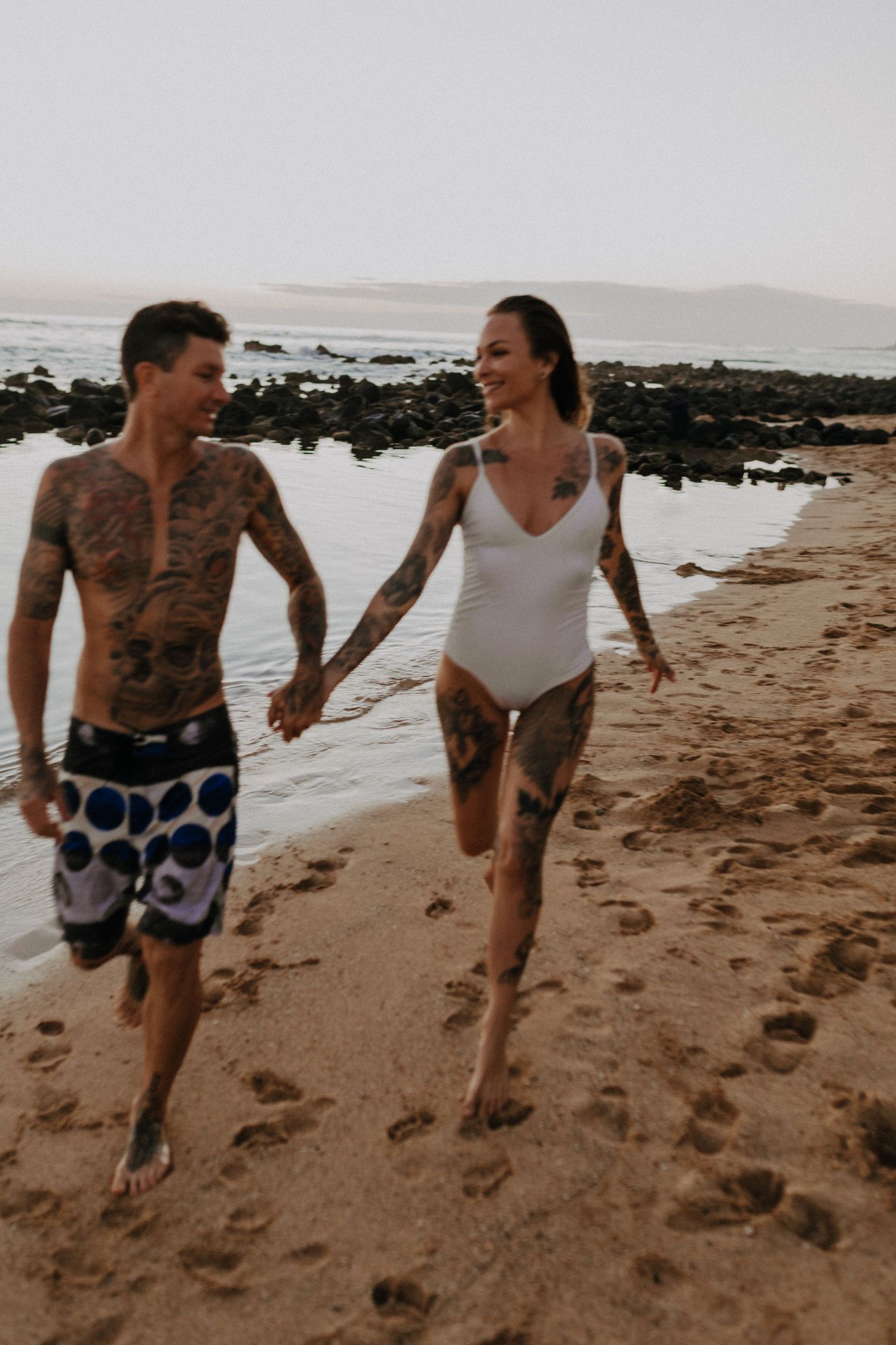 Playful couple runs and laughs together along a sandy beach with ocean waves in the background.