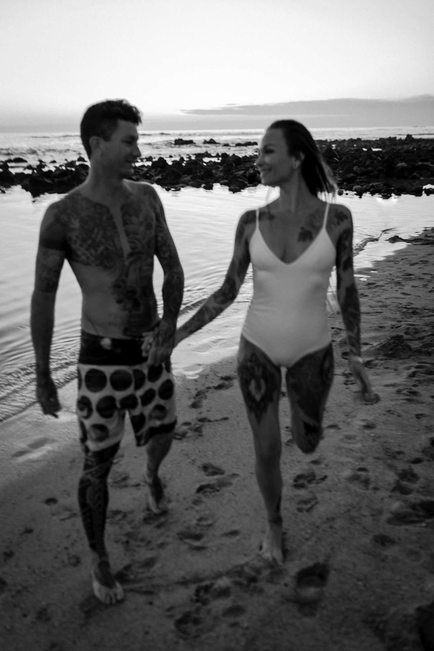 Couple walking together on a rocky beach shore with tattoos and swimwear in black and white.