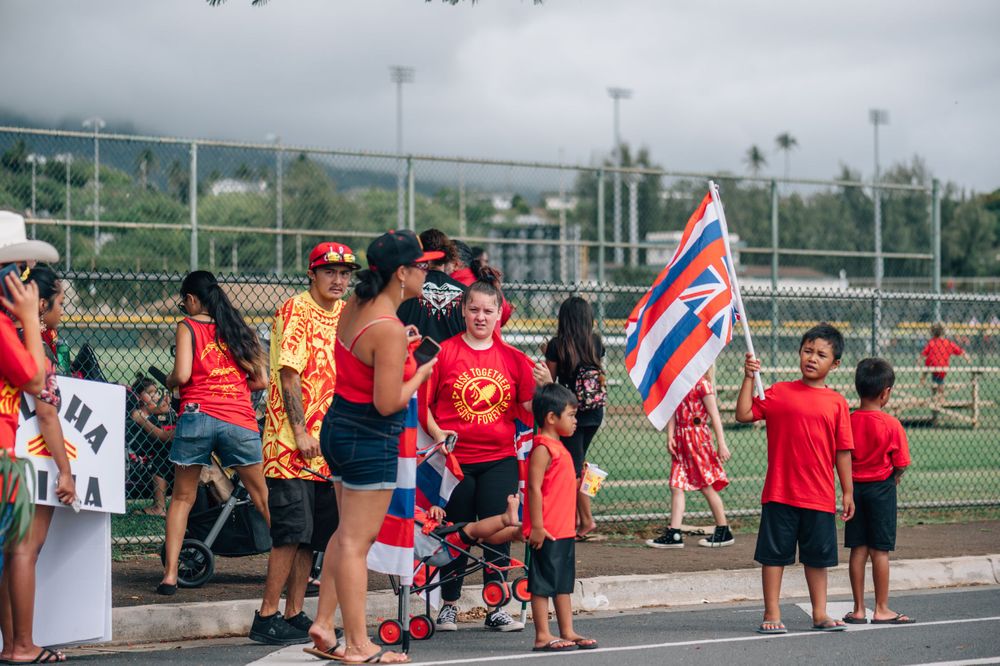 Spectators gather at a sports field with Hawaiian flags and red attire during a community event.