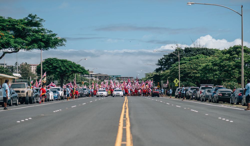 Wide view of a Hawaiian street lined with demonstrators and state flags.