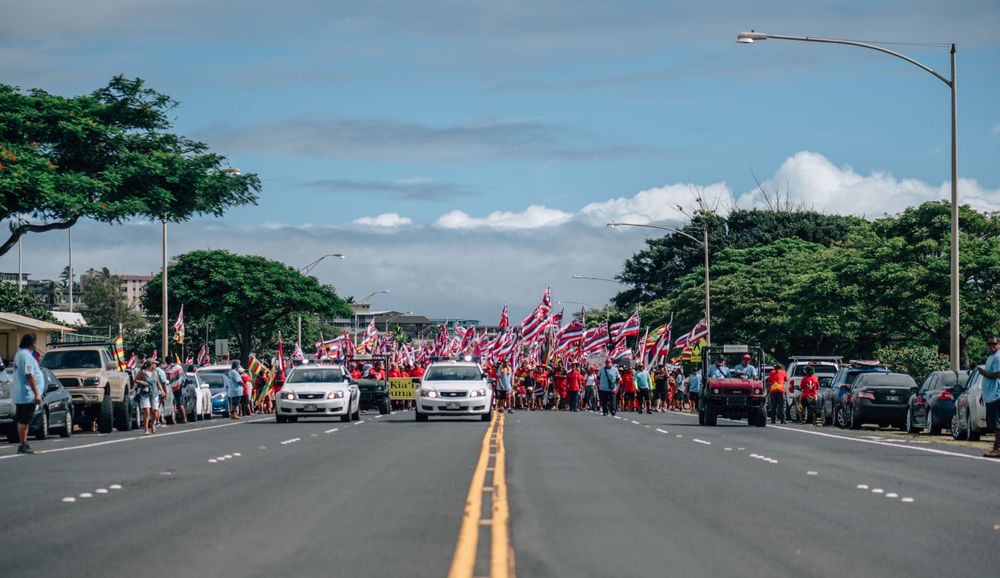 Procession of people carrying red flags marching between parked cars on a divided highway.