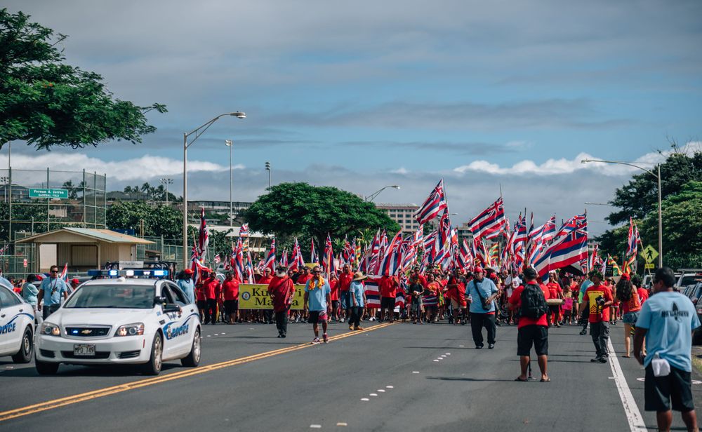 Large crowd marching down a street carrying Hawaiian state flags in a parade formation.