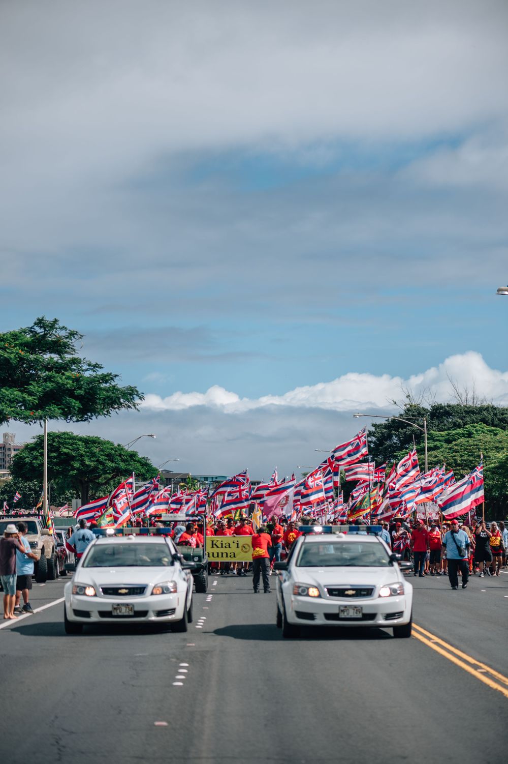 Two white cars lead a parade of flag-bearing marchers down a street lined with trees.