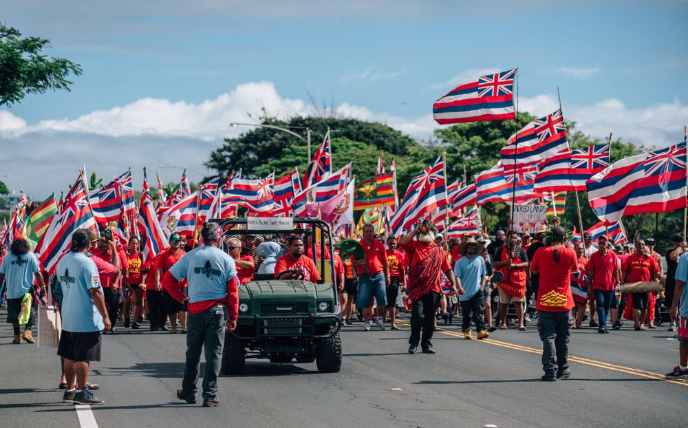 Crowd marches with Hawaiian state flags while wearing red shirts during a street demonstration.