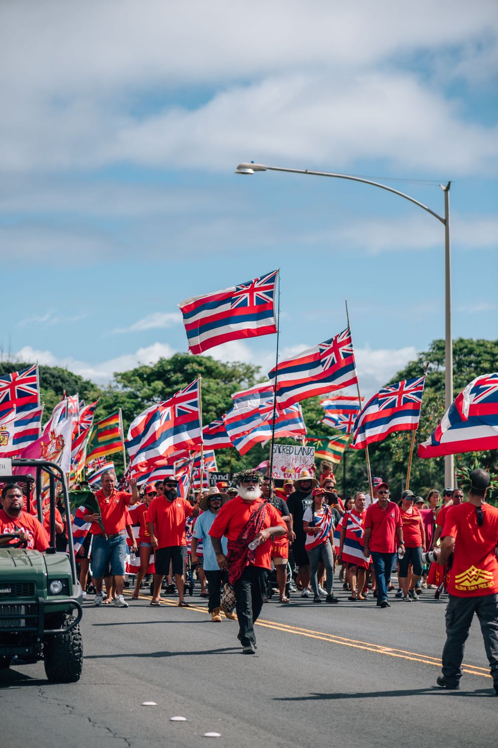Hawaiian flag bearers in red shirts march alongside a utility vehicle during a street parade.